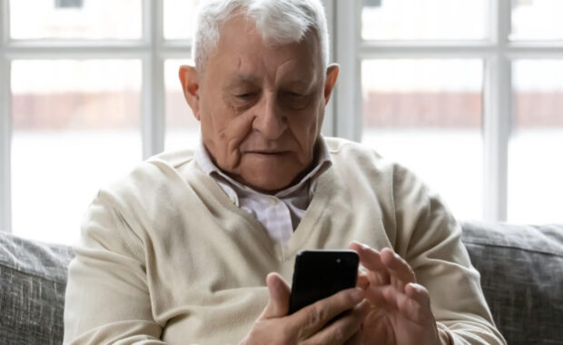 grey haired old man sit on sofa hold cellphone looking at screen
