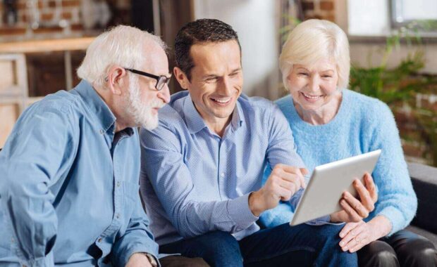 young man sitting on the couch between his senior parents and teaching them how to use a tablet