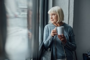 Lonely Senior Woman Holding Cup of Coffee Looking Through Window