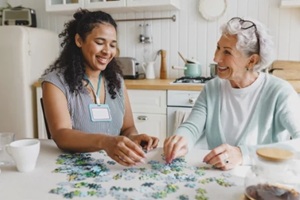 senior lady having fun plying puzzle game at kitchen table with black female volunteer, taking care of her as social support of retired persons